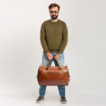 handsome confident bearded stylish man in sweatshirt with travel bag, wearing jeans and sunglasses isolated on white background